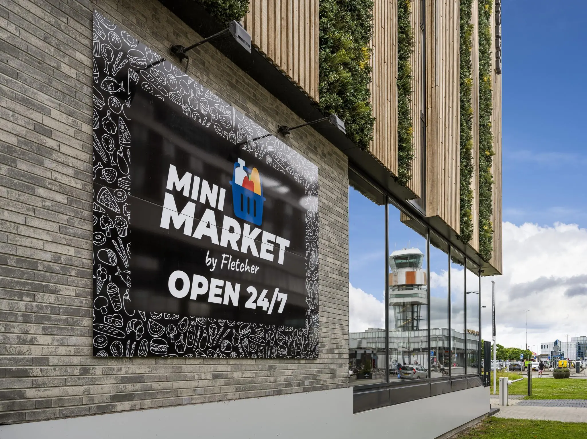 Gevel van een mini-market bij Fletcher Hotel op het Airportplein in Rotterdam, met zicht op de verkeerstoren van Rotterdam The Hague Airport.